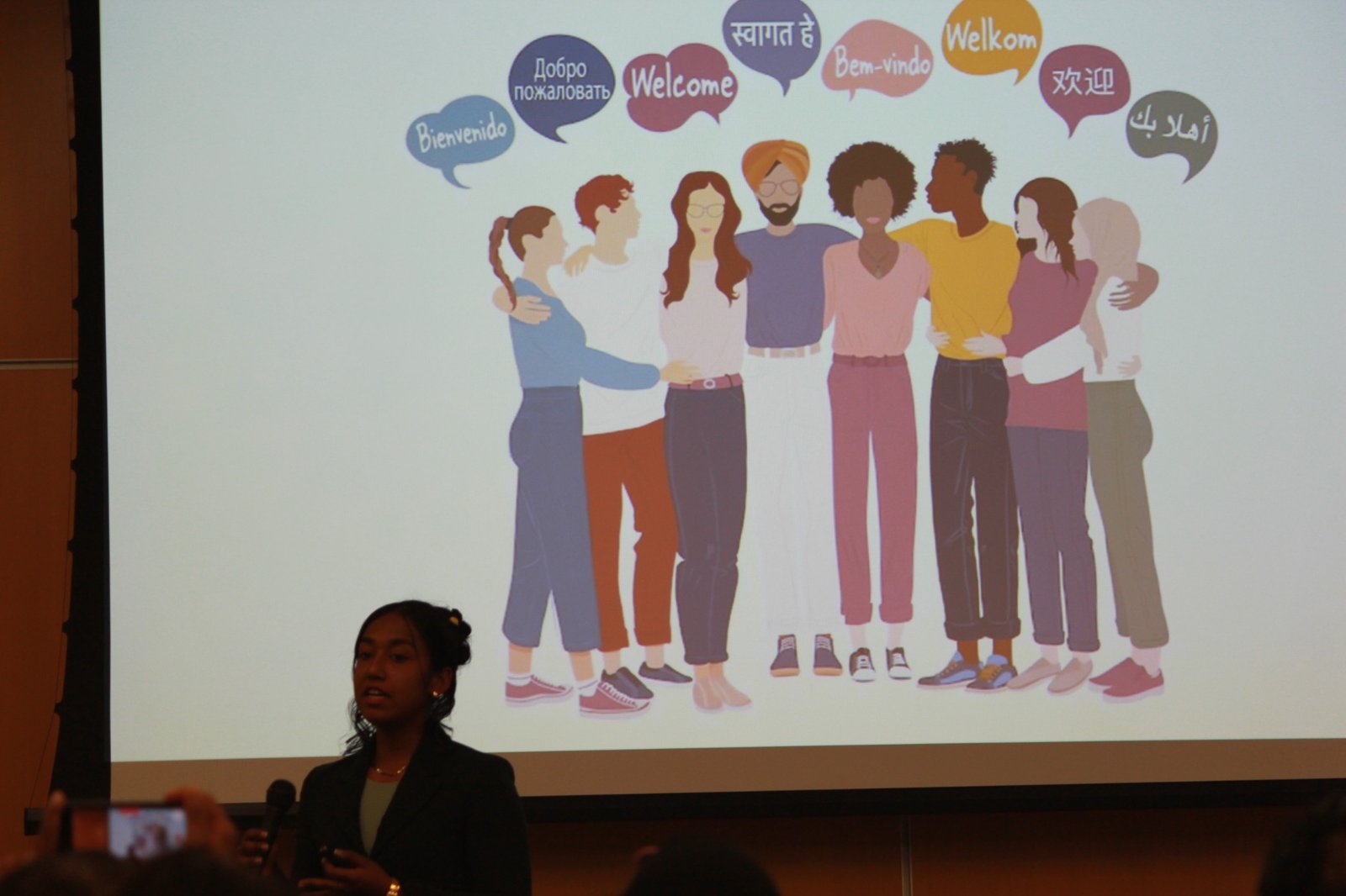 Ariana Rahman speaking in front of a multilingual welcome graphic